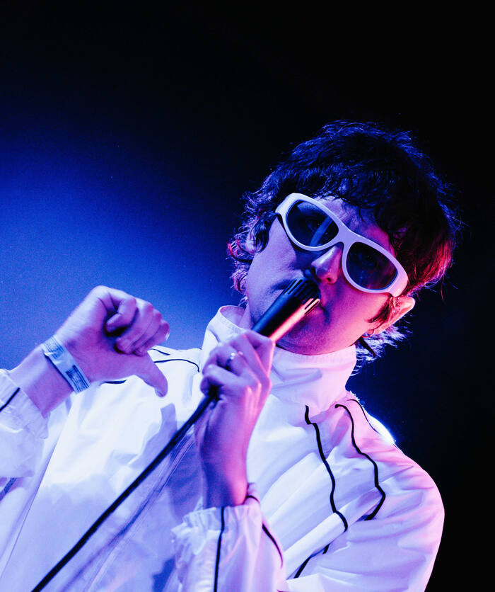 A singer in white sunglasses and a track jacket performing under blue and pink stage lights