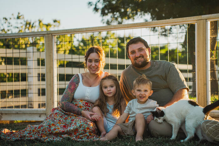 A family of four sitting on the grass at golden hour beside a wooden fence, with their cat